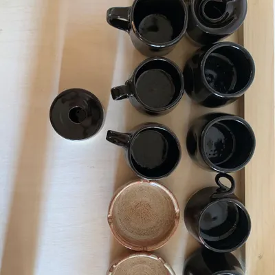 A collection of finished pieces, including glossy black mugs and rustic brown ashtrays. Each one is unique, with its own subtle variations in shape and glaze.