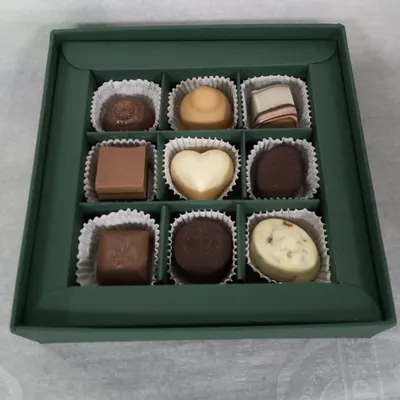 Inside a custom 9-piece box, you can see a variety of my handmade chocolates. This assortment includes a white chocolate heart, milk chocolate squares, and dark chocolate truffles, showcasing the different shapes and types I offer.