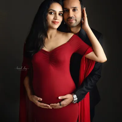 An elegant couple's maternity portrait against a dark grey backdrop. The woman's vibrant red gown and the intimate pose create a dramatic and romantic fine-art photograph.