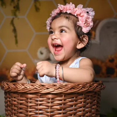 A heartwarming, happy moment captured during a photoshoot. The baby is wearing a floral crown that complements the sunflower and honeycomb backdrop, and is sitting in a rustic wicker basket.