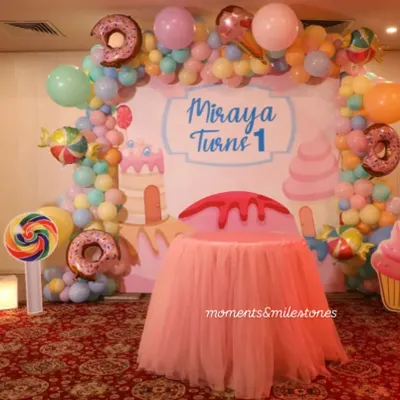 A sweet-themed cake-cutting area for Miraya's first birthday. The backdrop is surrounded by a pastel balloon arch with donut and candy-shaped foil balloons, and the cake table is dressed in a pink tutu skirt.