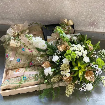 A closer look at one of our custom-curated gift hampers. This arrangement includes intricately embroidered shawls, complemented by fresh white and green florals and gold-accented foliage, all presented in a rustic wooden tray. We design each hamper to be a complete, luxurious gift for any special occasion.