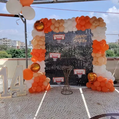 A basketball-themed decoration for a 14th birthday, set up on a sunny terrace. This features a balloon arch in orange and white, a silver sequin backdrop with basketball hoop details, and large light-up marquee numbers for the age.