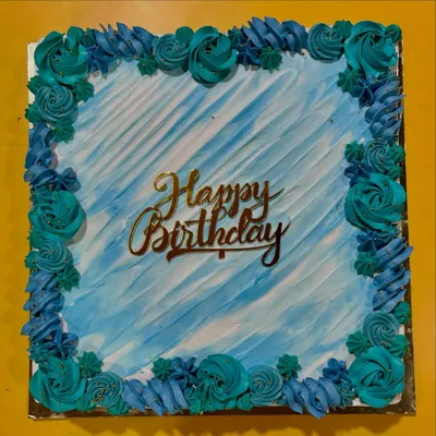 A beautiful square birthday cake with a watercolor-effect blue frosting. The border is decorated with hand-piped buttercream rosettes in shades of teal and blue.