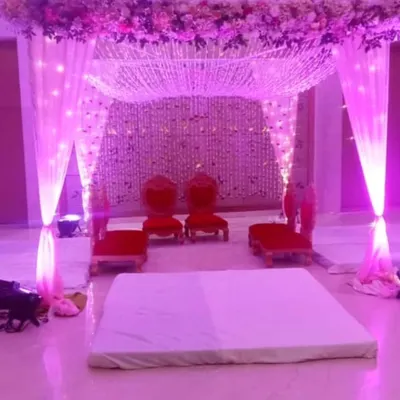 An indoor mandap setup illuminated with beautiful purple lighting. The ceiling is decorated with a web of floral strings, and the seating is arranged for the wedding rituals.