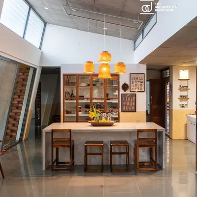 The central dining island at 'Aamra', crafted from polished cement and paired with simple wooden stools. The space is illuminated by natural light from a clerestory window, highlighting the raw, unplastered textures of the home.