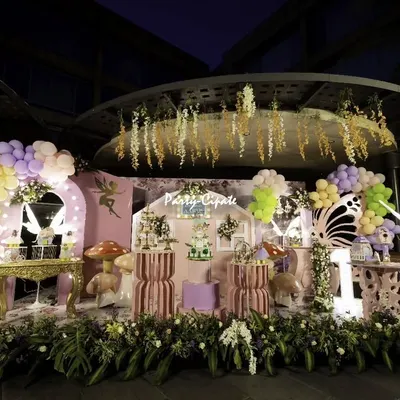 A full view of a fairy and butterfly themed birthday party at dusk. The setup includes a central stage designed like a cottage, flanked by balloon arches, illuminated butterfly wings, and lush floral arrangements at the base.
