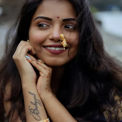A close-up portrait capturing a joyful, natural smile. The focus is on her expressive face, traditional nath (nose ring), and the 'Babygal' tattoo, showing how modern style and tradition can blend beautifully in a single frame.