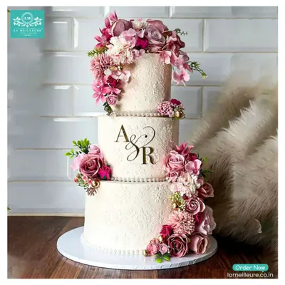 This three-tiered wedding cake features a rustic textured buttercream finish and is personalized with the couple's initials, 'A & R'. The design is elevated by a cascading arrangement of fresh flowers in shades of mauve and deep pink.
