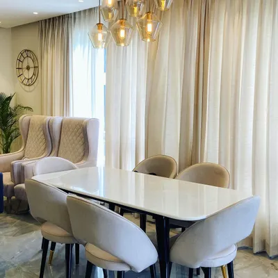 The centerpiece of this dining room in Noida is the stunning cluster of glass pendant lights, which casts a warm glow over the marble-top table. The fully upholstered, curved-back chairs provide exceptional comfort, while the sheer curtains diffuse natural light to create a soft and elegant atmosphere.