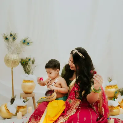 A beautiful 'Maiya aur Kanha' moment from a Janmashtami themed shoot. The mother and son are surrounded by peacock feathers and pots, creating a rich, festive scene.