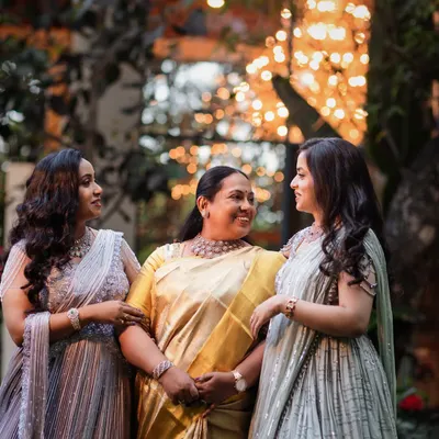 A heartwarming moment between a mother and her two daughters, all dressed in custom Needle Eye creations. The daughters' pastel lehengas beautifully complement their mother's elegant golden saree.