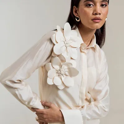 Minimalist structure, maximalist detail. This ivory shirt is elevated with three-dimensional floral appliqués on the shoulder, a true statement of wearable art.