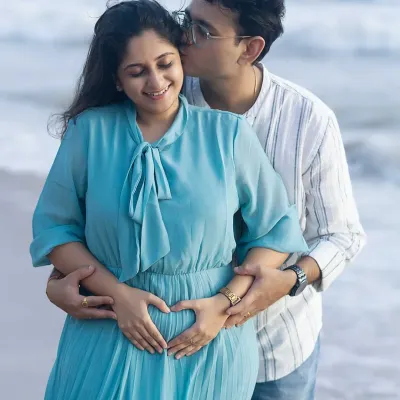 A truly heartwarming moment. The dad-to-be kisses his partner's cheek as they both gently form a heart with their hands on the baby bump, with the ocean waves in the background.