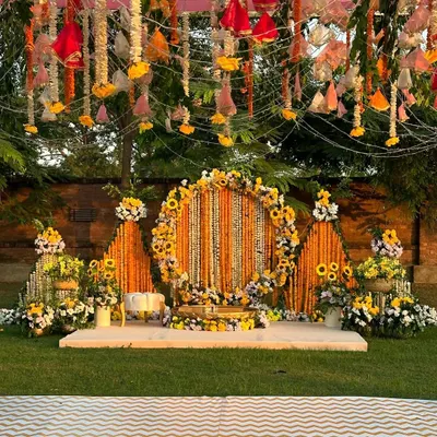This outdoor Haldi setup is a celebration of sunshine and tradition. A circular stage is framed by pyramids of marigold garlands, creating a vibrant and joyful space for the ceremony.