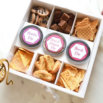 This is our Grazing Box, a perfect Rakhi hamper for sharing. It includes a mix of sweet and savory treats like choco-chip cookies, brownie bites, assorted crackers, and three of our signature dips: Nutella, Cheese & Jalapeno, and Biscoff.