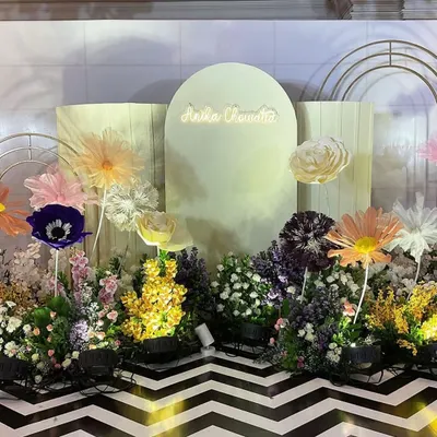 A whimsical garden stage for a cradle ceremony, featuring a neon sign with the baby's name, oversized paper flowers, and a bed of colorful wildflowers. The chevron floor adds a modern, graphic touch.
