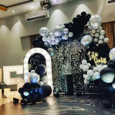 A stylish 25th birthday party setup with a black and silver theme. This decor features a silver shimmer wall, a "Let's Party" neon sign, large '25' marquee numbers, and an elaborate balloon arrangement.