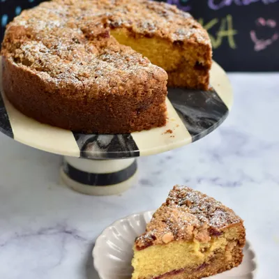 Our Plum & Almond Crumble Tea Cake, with a slice ready to be enjoyed. This comforting cake has a layer of plum jam inside and a crunchy almond crumble on top, perfect for a rainy afternoon with chai.