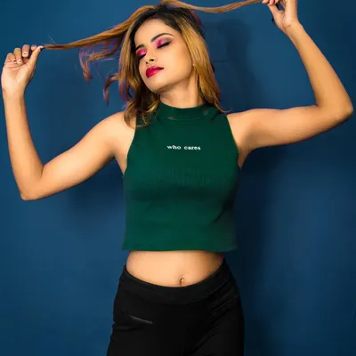 A playful and confident studio portrait. The model, wearing a green crop top, plays with her hair against a solid blue background, creating a vibrant and energetic fashion shot.