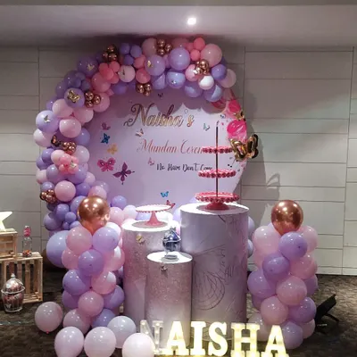 A lovely setup for "Naisha's Mundan Ceremony" with a butterfly theme. The design features a large circular backdrop, a balloon arch in purple and pink, and illuminated name letters, creating a perfect photo opportunity.