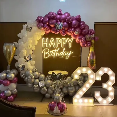 A complete birthday party setup for a 23rd celebration, featuring a bright 'Happy Birthday' neon sign and illuminated marquee numbers. The balloon arch uses a mix of purple, silver, and white, with butterfly props adding a whimsical touch.