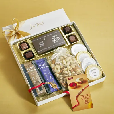 Our white-and-gold Rakhi hamper combines assorted chocolates, dry fruits, Hot Chocolate Mix and a card beside a red Rakhi.