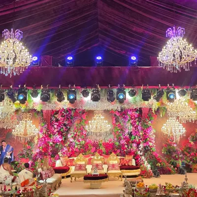 A wide view of a spectacular wedding stage under a tent, illuminated by multiple crystal chandeliers. The entire backdrop is a dense wall of red and pink flowers, creating an opulent and unforgettable setting.