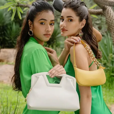 A close-up from the Sarjaa sustainable handbag campaign. This shot highlights the interaction between the models and the products, showcasing two different bag styles in a lush, green environment.