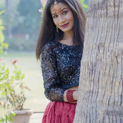 A smiling, candid moment from an outdoor ethnic fashion shoot. The model is peeking from behind a tree, creating a playful yet professional portrait.