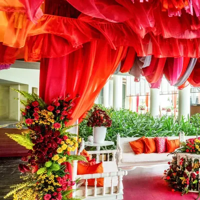 A vibrant Mehendi setting with a canopy of layered red, pink, and orange drapes. The seating area is framed by lush floral pillars, creating a cozy and festive atmosphere.