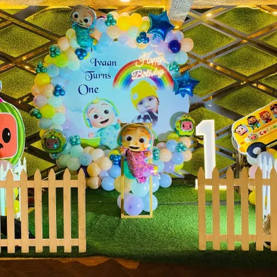A vibrant Cocomelon theme for a first birthday party. This setup features a custom backdrop with characters, a colorful balloon arrangement, character cutouts, and a picket fence prop to create a fun and immersive experience.