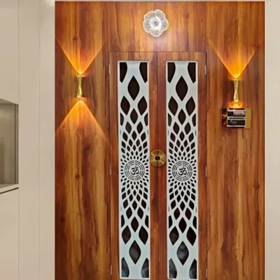 The entrance to a home mandir, featuring wooden doors with beautiful white jaali work. The warm wall lights on either side create a welcoming and divine ambiance right at the doorway.