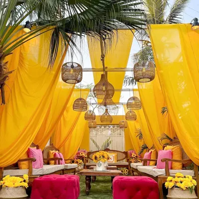 A vibrant Haldi or Roka setup featuring bright yellow drapes, cane furniture, and pops of fuchsia. The combination of sunflowers and bohemian-style lanterns creates a cheerful and relaxed vibe.