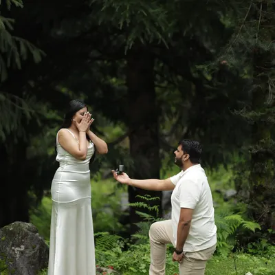 A full view of the surprise proposal, with red petals scattered on the forest floor. This shot captures the entire scene, from his hopeful expression to her genuine shock.