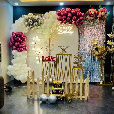 A popular birthday decoration featuring a white, silver, and magenta balloon arch against a shimmering backdrop. The neon "Happy Birthday" sign and decorative pedestals complete the look for a festive party venue.