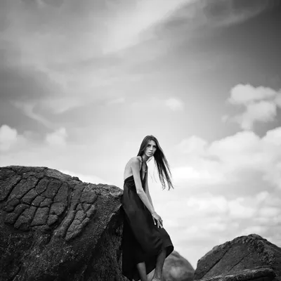 This black and white composition emphasizes the contrast between the dark rocks and the bright, textured sky. The model's pose creates a strong, graphic silhouette.