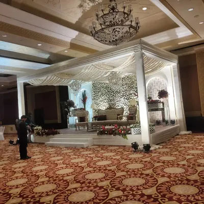 A luxurious indoor wedding stage featuring a white mandap structure against a full wall of white flowers. The setup is completed with traditional seating and enhanced by the hall's ornate crystal chandelier.