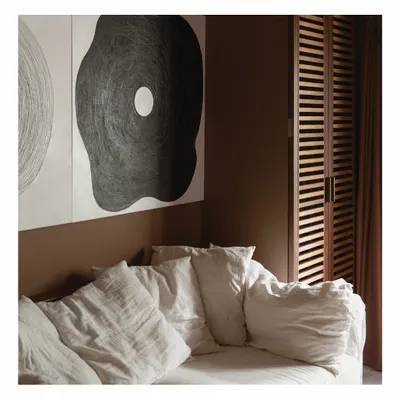 A comfortable slipcover sofa sits against a warm, earthy brown wall, creating a cozy corner for relaxation. The large, abstract artwork and slatted wooden screen add layers of texture and interest, defining the room's calm and artistic character.