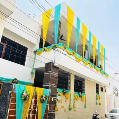 A house exterior decorated with bright yellow and turquoise drapes for a pre-wedding ceremony. The combination of colors on the balcony and gate, accented with small floral bouquets, creates a cheerful and festive look.