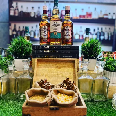 The heart of the Whisky and Cigar bar. Premium bottles like The Glenlivet and Chivas Regal are elegantly displayed on a stack of books, surrounded by tasting elements like cinnamon sticks and grains in rustic burlap sacks.