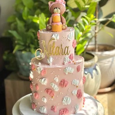 A little pink dream for a sweet one-year milestone. This two-tier cake features palette-knife florals, a custom gold name topper, and an adorable, hand-crafted teddy bear holding her own mini cake.