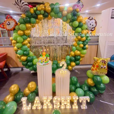A fun jungle safari theme for a second birthday. This setup features a circular balloon arch in shades of green and gold, a silver fringe backdrop, animal character cutouts like a monkey and giraffe, and light-up marquee letters.