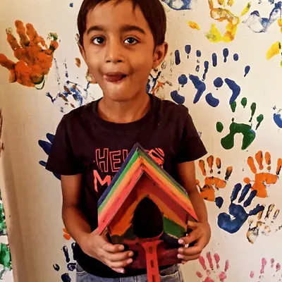 A proud young builder shows off the rainbow-colored birdhouse he made, standing in front of our signature handprint wall.
