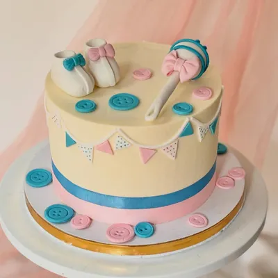 A classic baby shower cake with a gender reveal theme. This cake is decorated with pink and blue buttons, a bunting banner, and adorable baby booties and a rattle on top.