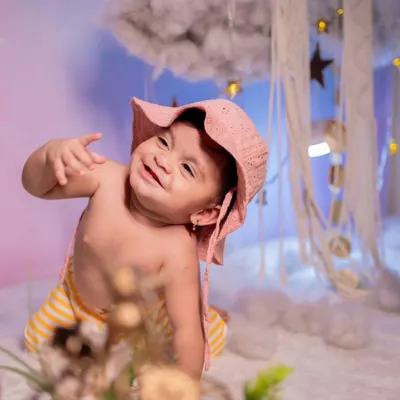 An adorable and playful shot of a baby with a huge smile, wearing a cute pink hat. The dreamy background with clouds and stars adds a whimsical touch to this indoor baby shoot.