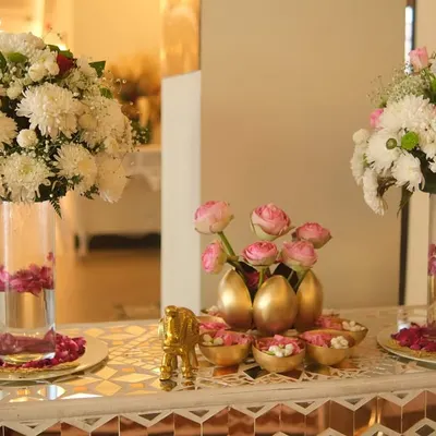 An elegant welcome table setup featuring tall glass vases with large bouquets of white chrysanthemums. The centerpiece includes pink lotus buds in golden egg-shaped vases.