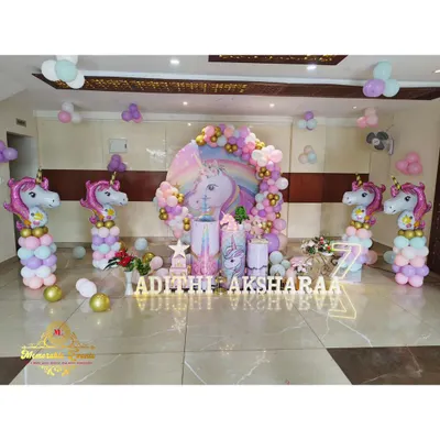 A spectacular unicorn-themed party setup with a large unicorn backdrop, pastel balloon arch, and unicorn-shaped balloon columns. The floor is decorated with scattered balloons and marquee letters for the names.