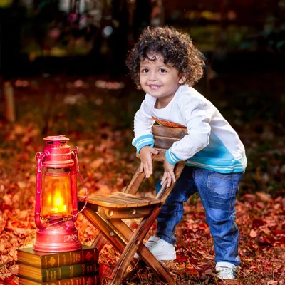 A toddler boy with amazing curly hair leans on a chair in a forest setting, surrounded by autumn leaves and a glowing lantern. The warm, magical light creates a stunning outdoor portrait.