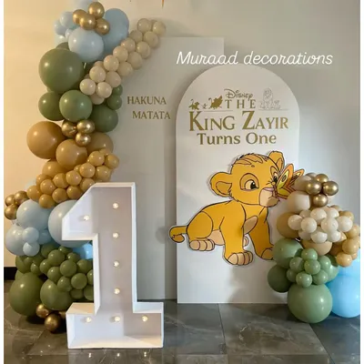 A 'Hakuna Matata' first birthday featuring a Lion King theme. This setup includes a large light-up number '1', a custom arched backdrop with Simba, and a balloon garland in safari-inspired colors like olive green and tan.